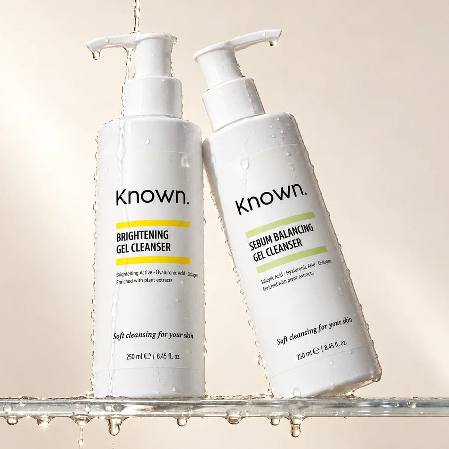 Two white pump bottles of Known skincare cleansers on a glass surface with water droplets and dripping water, against a warm beige background. One bottle is labeled Brightening Gel Cleanser and the other is labeled Serum Balancing Gel Cleanser.
